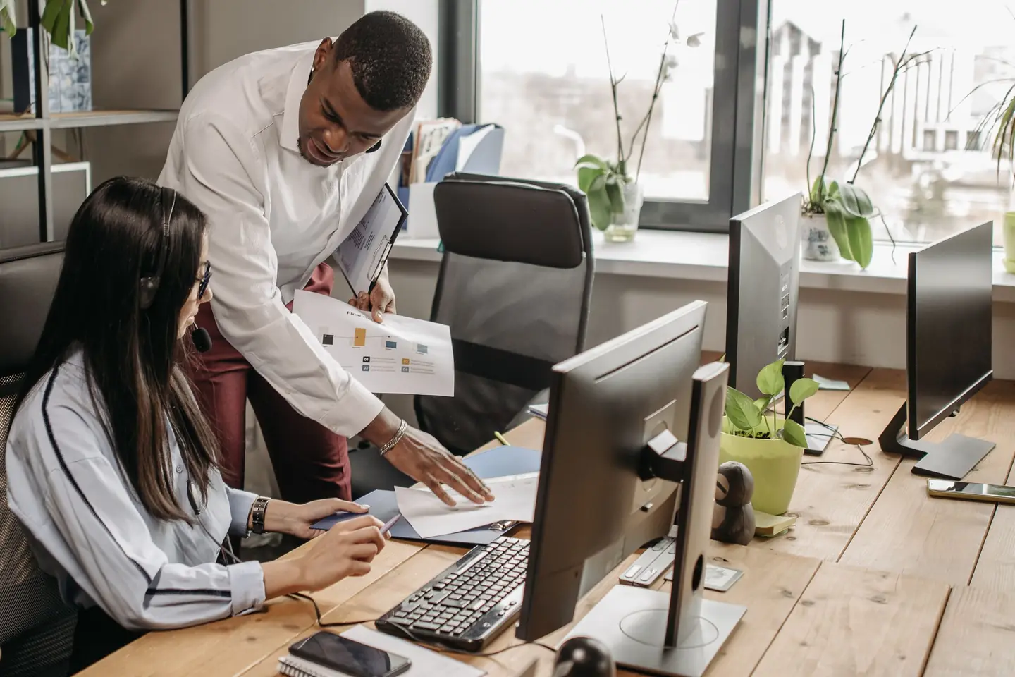 A man leaning over a desk to help a coworker at her computer in a bright office
